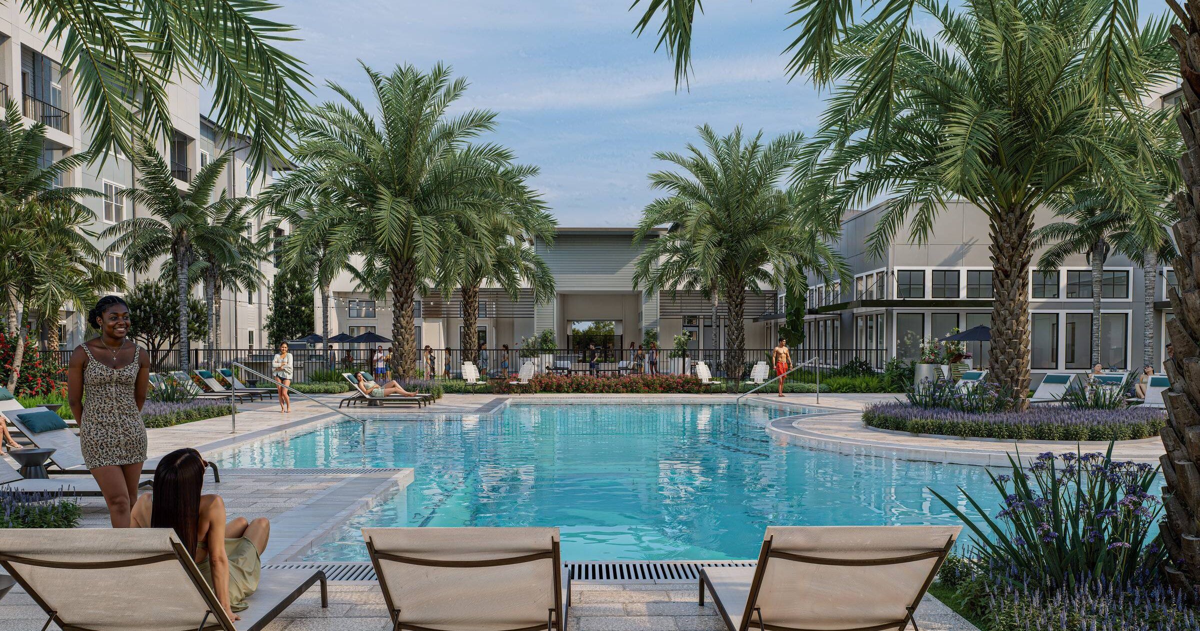 People relax by a large outdoor pool surrounded by palm trees, lounge chairs, and apartment buildings.